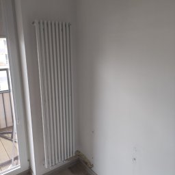 Ark - Biały grzejnik panelowy zamontowany przy ścianie w rogu pokoju, obok okna balkonowego. Widoczne ślady po starym grzejniku i pianka montażowa w szczelinie przy podłodze.