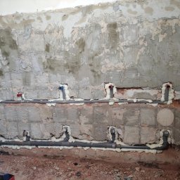 Ark - Szare rury instalacji hydraulicznej zamontowane w ścianie z surowego betonu, zabezpieczone pianką montażową. Widoczne fragmenty instalacji z czerwonymi i niebieskimi końcówkami.