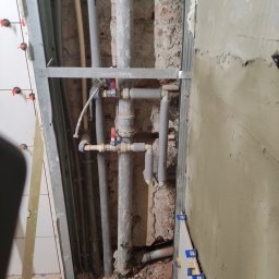 Ark - Montaż instalacji wodnej z rurami w szarej izolacji, widoczne zawory i fragment ściany z płytkami. Prace hydrauliczne w trakcie remontu łazienki.