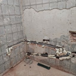 Ark - Szare rury hydrauliczne, zabezpieczone pianką montażową, biegną wzdłuż ściany z resztkami płytek w trakcie remontu łazienki. Widoczny fragment podłogi i ściany.
