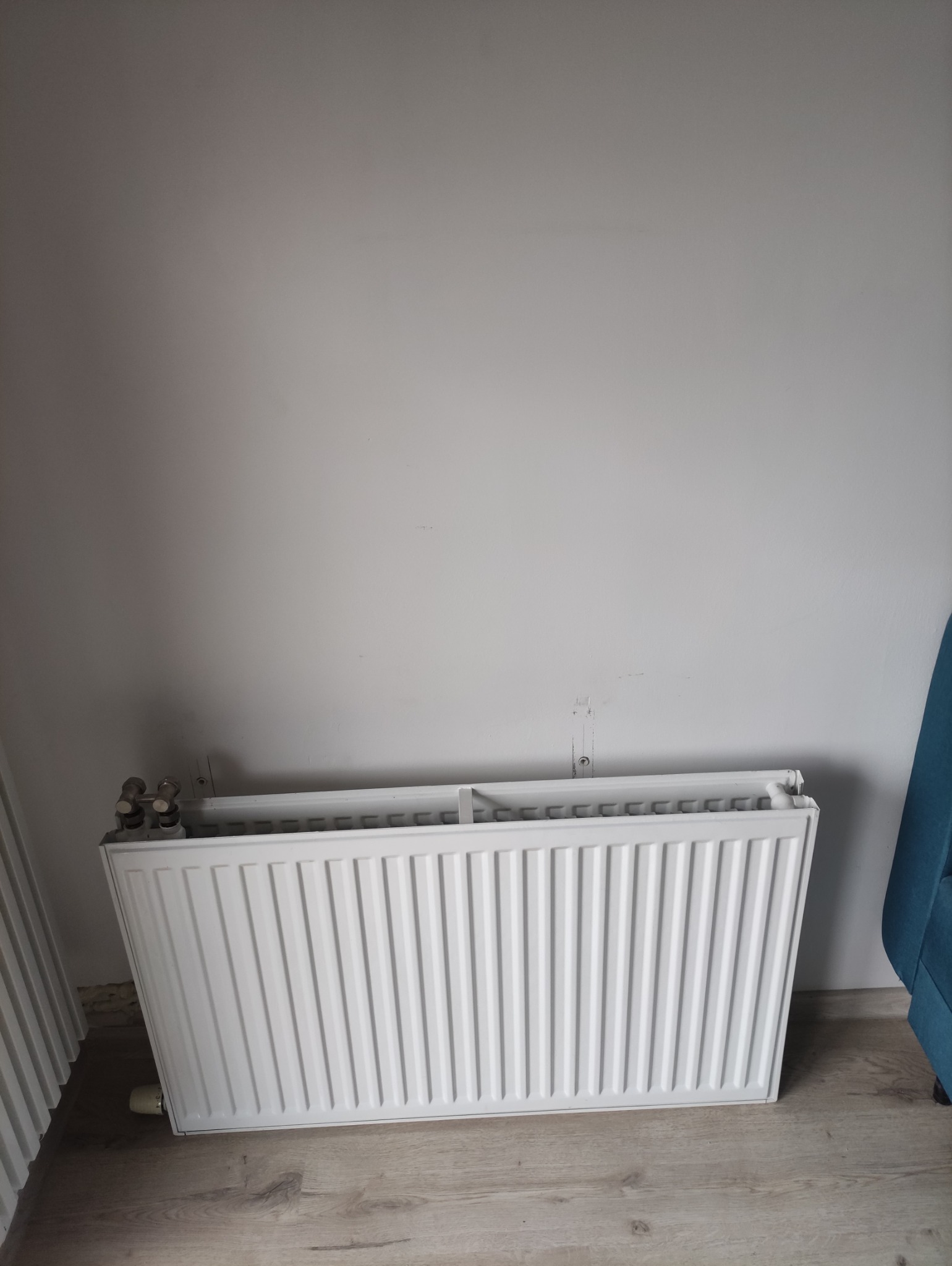Biały grzejnik panelowy, częściowo odsłonięty, oparty o ścianę na drewnianej podłodze. Widoczne ślady po poprzednim montażu na ścianie.
