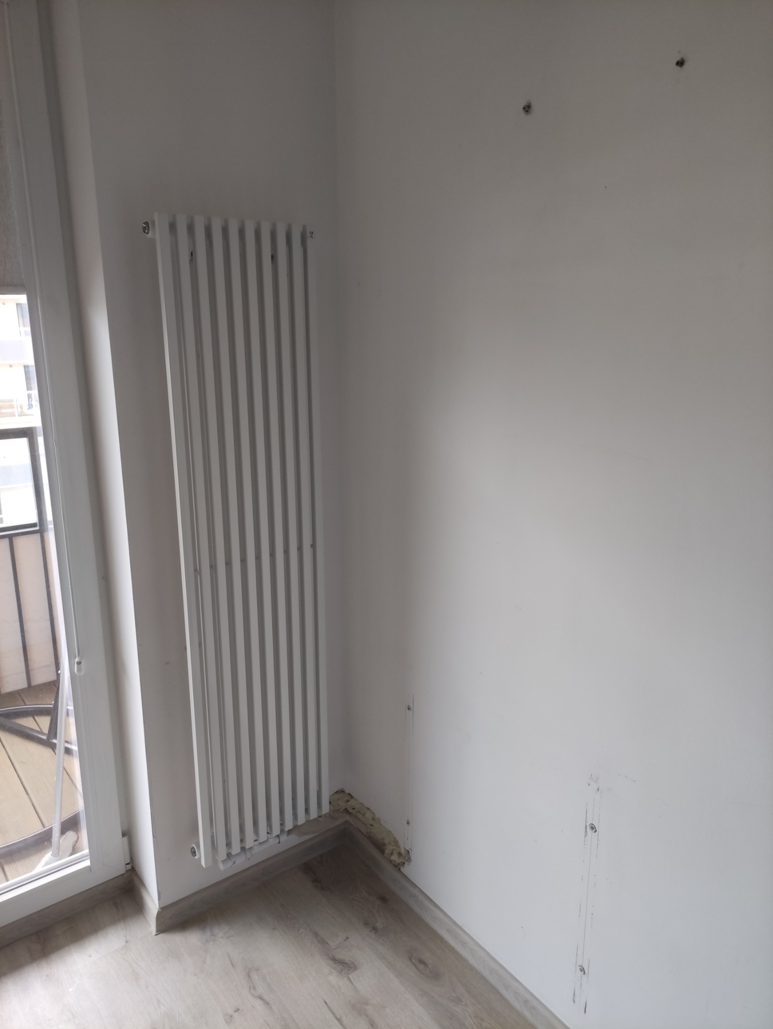 Biały grzejnik panelowy zamontowany przy oknie balkonowym na tle białej ściany z widocznymi śladami po instalacji. Jasne panele podłogowe.
