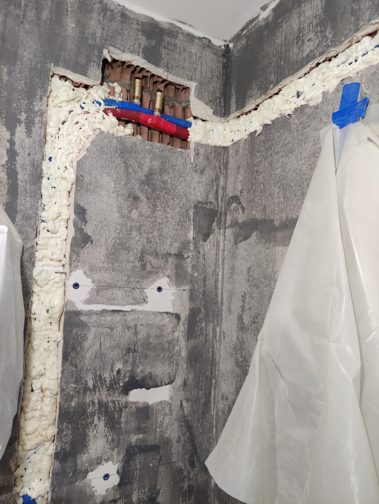 Ściana w trakcie prac hydraulicznych: widoczne rury (czerwona i niebieska) w otworze, uszczelnione pianką montażową. Część ściany zabezpieczona folią malarską.