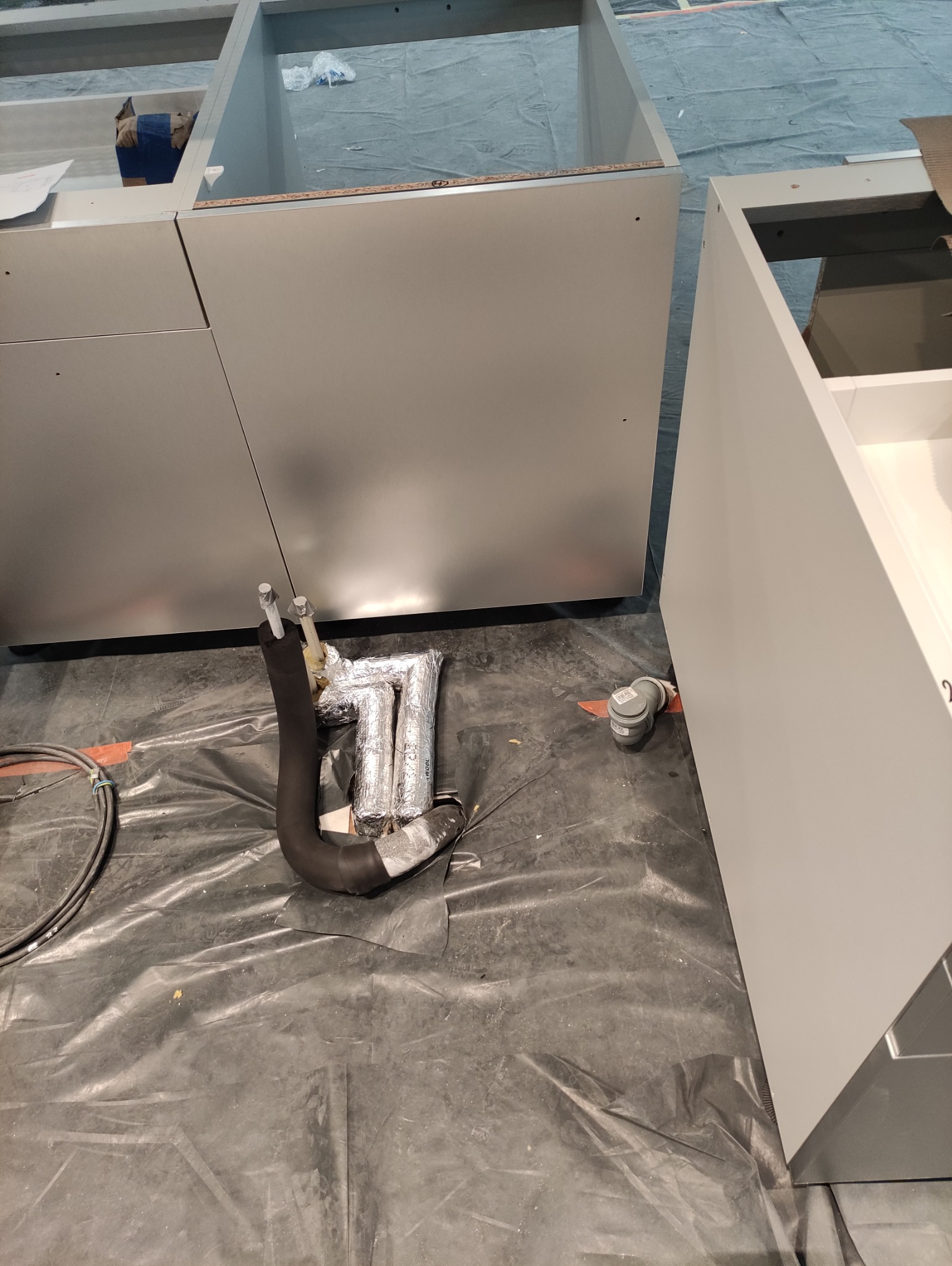 Fragment instalacji hydraulicznej z rurami w izolacji, widoczne elementy montażowe i fragmenty szafek kuchennych w trakcie montażu na folii ochronnej.