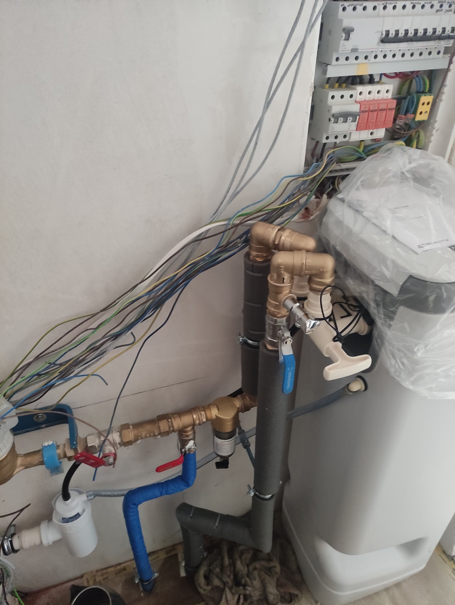 Instalacja wodna z filtrem i zmiękczaczem wody, widoczne miedziane rury, szare izolacje, biały zbiornik i rozdzielnia elektryczna na ścianie.