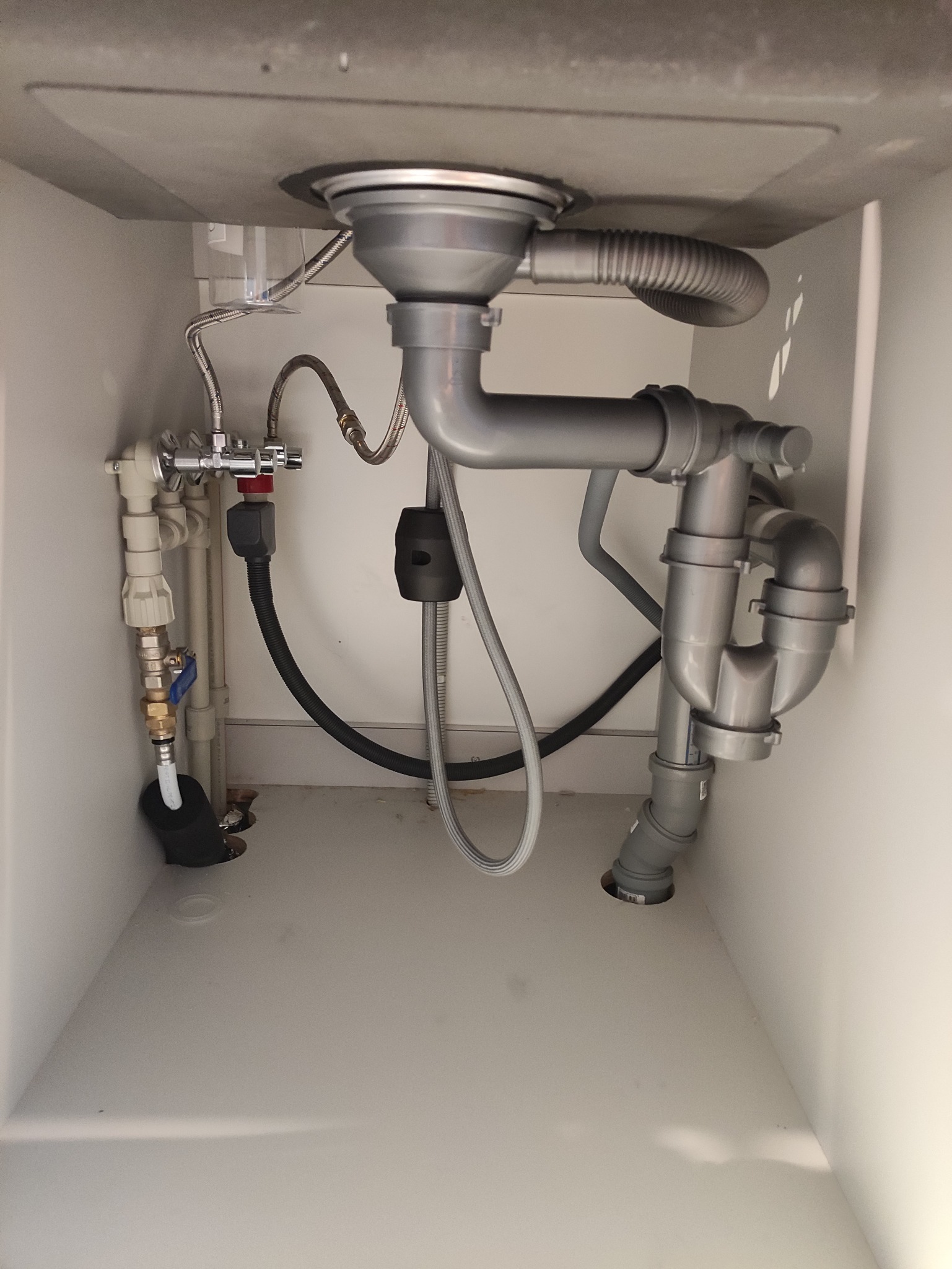 Szare rury odpływowe i zawory pod zlewem w białej szafce. Widoczne złącza, wężyki i syfon. Ujęcie z dołu, ukazujące instalację hydrauliczną.