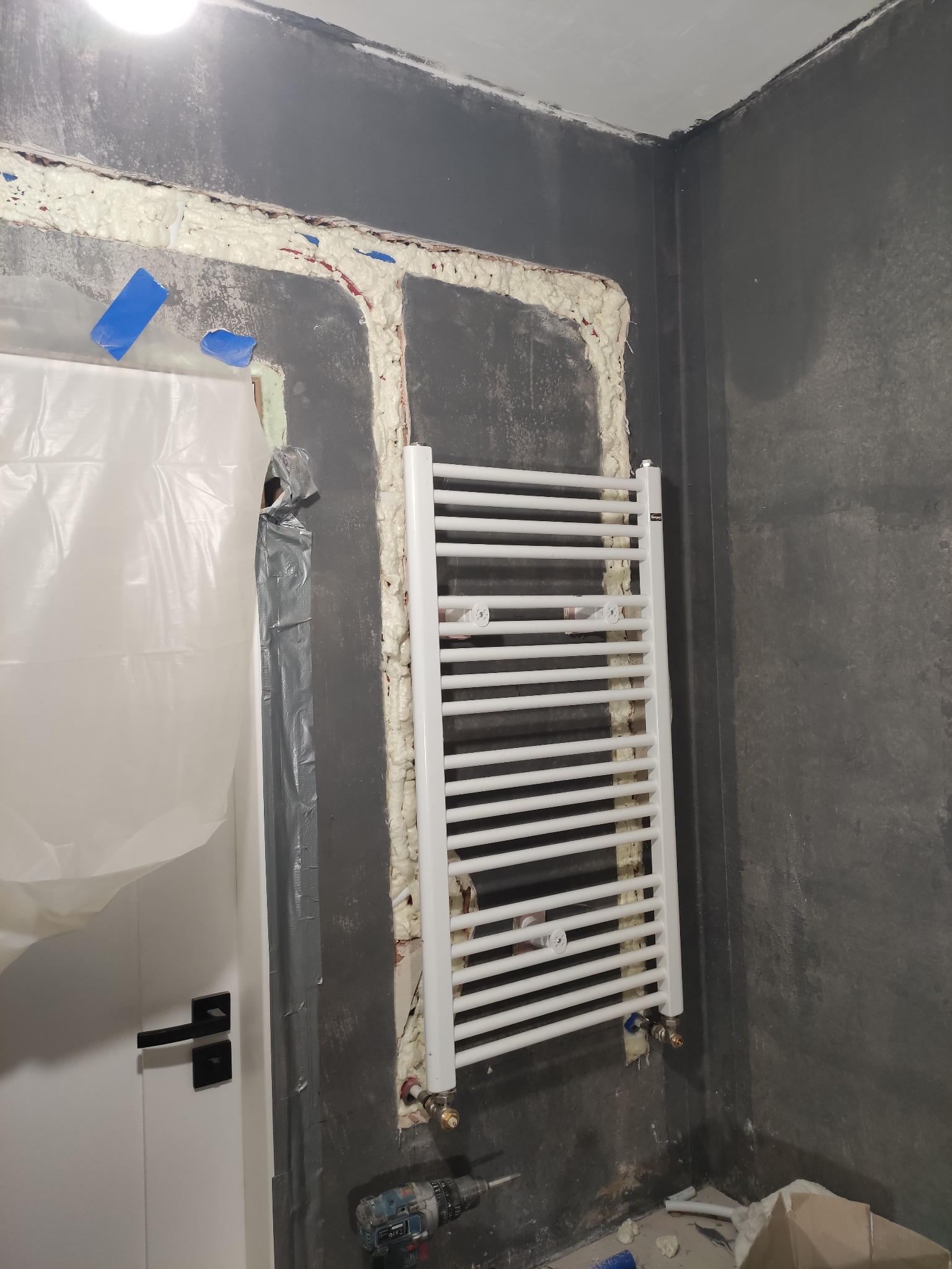 Biały grzejnik łazienkowy zamontowany na szarej ścianie, z widoczną pianką montażową wokół rur. W tle zakryte folią drzwi i wiertarka na podłodze.
