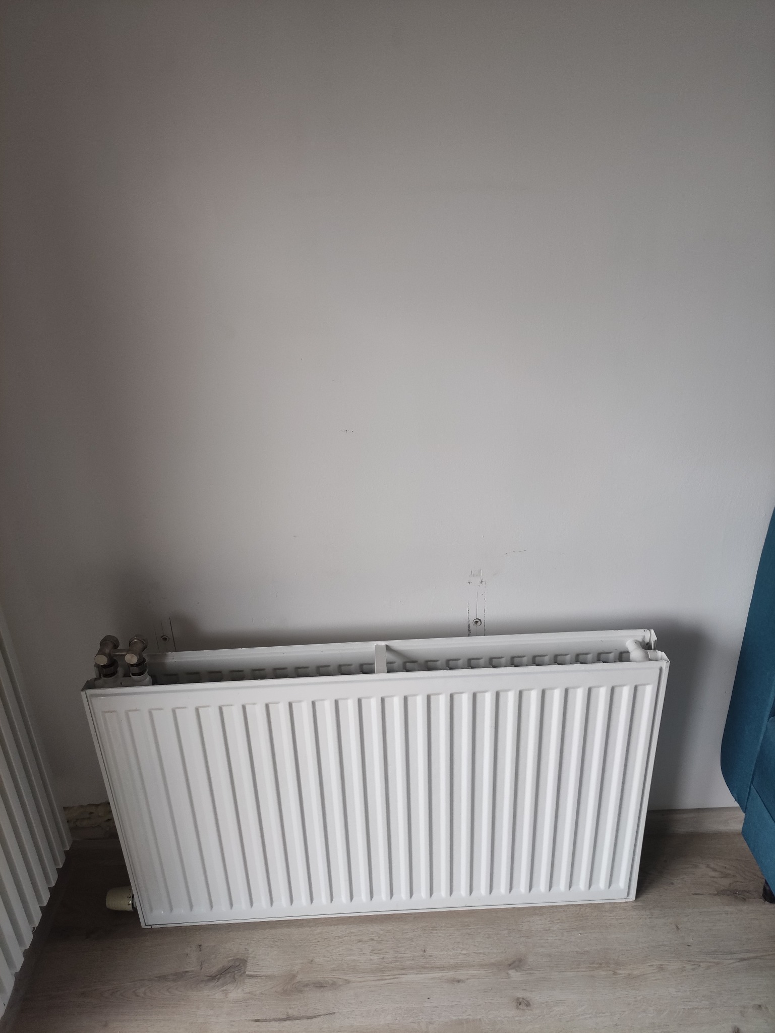 Biały grzejnik panelowy, zamontowany na ścianie we Wrocławiu. Widoczne ślady po starym montażu. Ujęcie z dołu, fragment niebieskiego fotela po prawej.