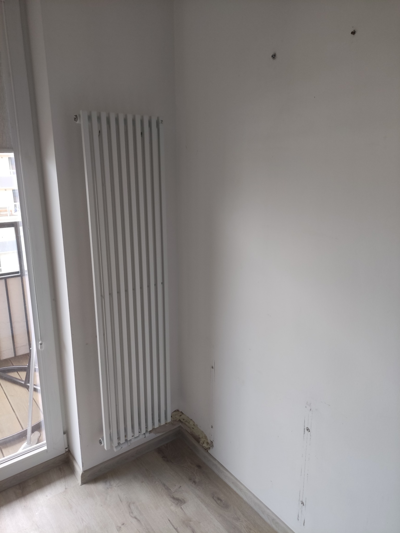 Biały grzejnik panelowy zamontowany przy ścianie w rogu pokoju, obok okna balkonowego. Widoczne ślady po starym grzejniku i pianka montażowa w szczelinie przy podłodze.