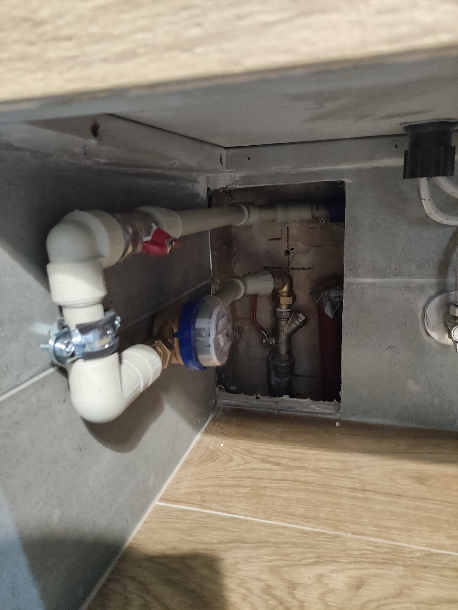 Zamontowany wodomierz z zaworem odcinającym i rurami z tworzywa w zabudowie pod umywalką. Szare płytki i drewnopodobna podłoga.