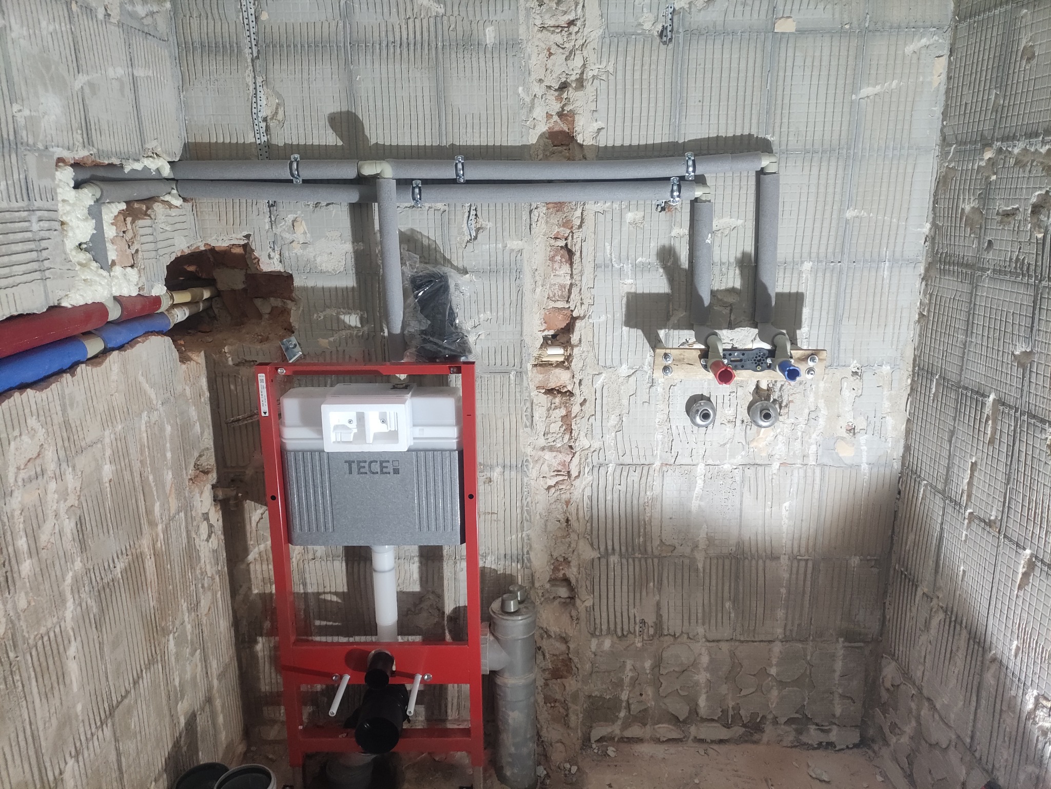 Stelaż podtynkowy WC marki TECE z szarym zbiornikiem, widoczne rury w izolacji i przyłącza na tle surowej ściany w trakcie prac hydraulicznych.