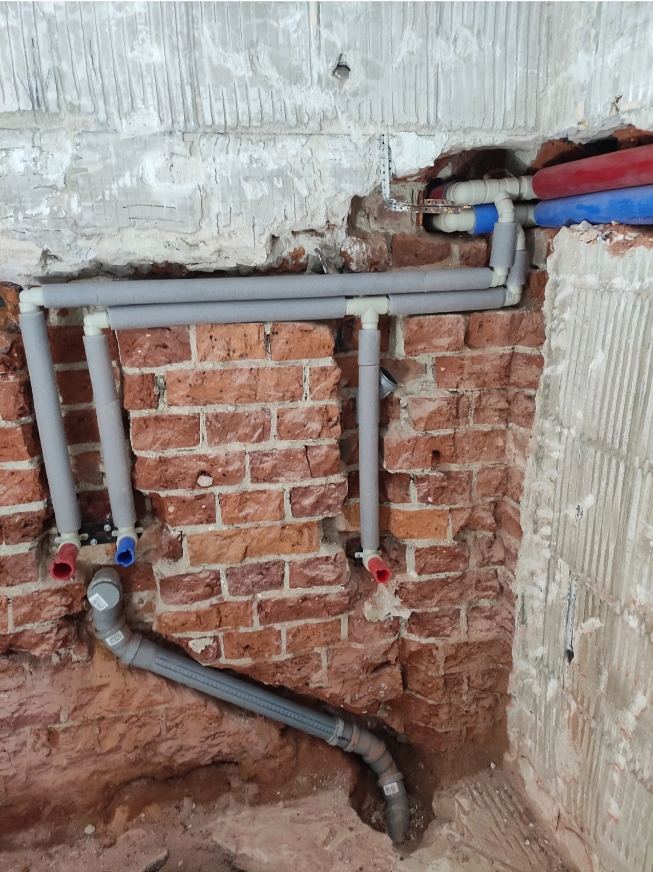 Fragment ściany z odsłoniętą czerwoną cegłą, na której zamontowano szare, zaizolowane rury instalacji wodnej z czerwonymi i niebieskimi końcówkami.