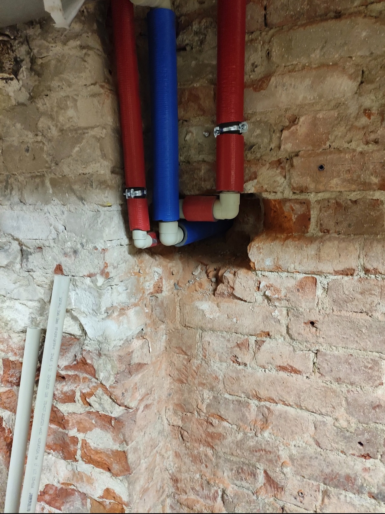 Instalacja wodna z rur PEX w kolorze czerwonym i niebieskim, zamocowana do ściany z cegły. Widoczne złączki i obejmy mocujące. Fragment rurki PCV leży przy ścianie.
