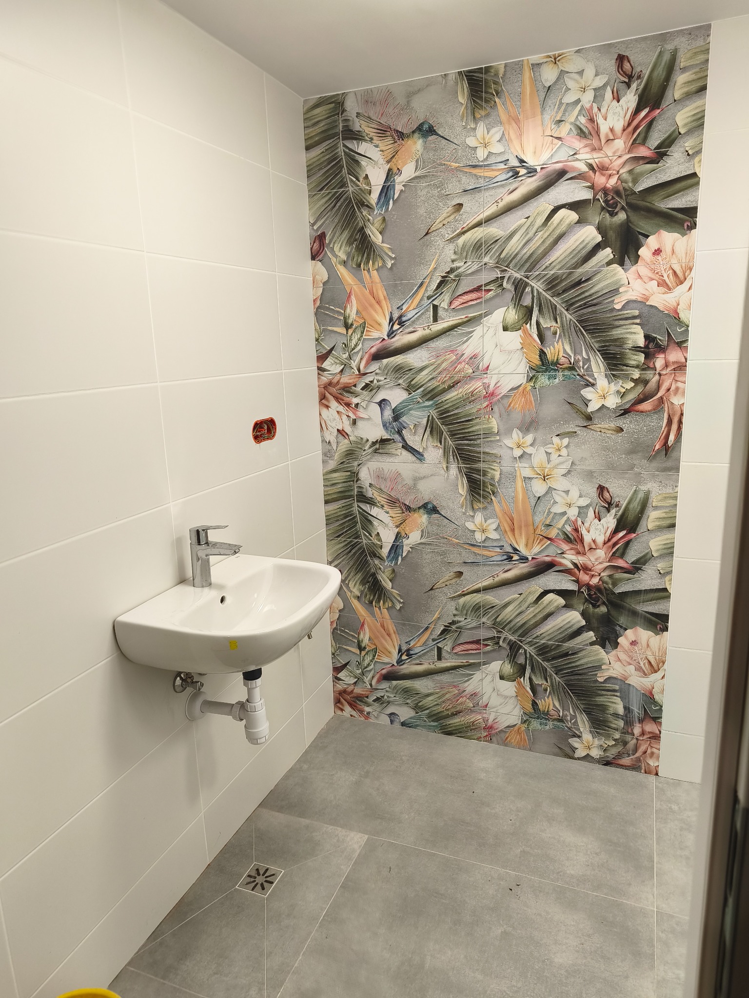 Biała umywalka z chromowaną baterią na tle ściany z białych płytek i dekoracyjnej mozaiki z motywem tropikalnych kwiatów i ptaków w wykończonej łazience.