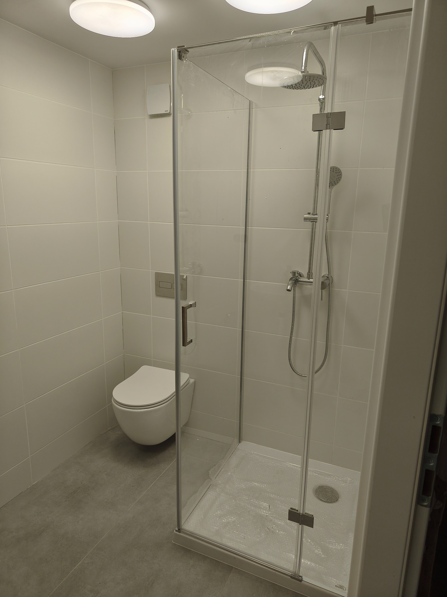 Nowoczesna kabina prysznicowa z deszczownicą i szklanymi drzwiami, widoczna toaleta podtynkowa, białe płytki na ścianach, szara podłoga. Minimalistyczny design łazienki.