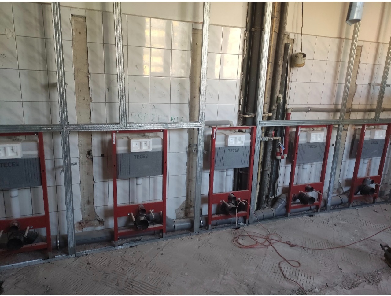 Montaż stelaży podtynkowych TECE w łazience z widocznymi rurami i instalacją. Ściany w trakcie wykończenia, widoczne płytki i metalowe profile.