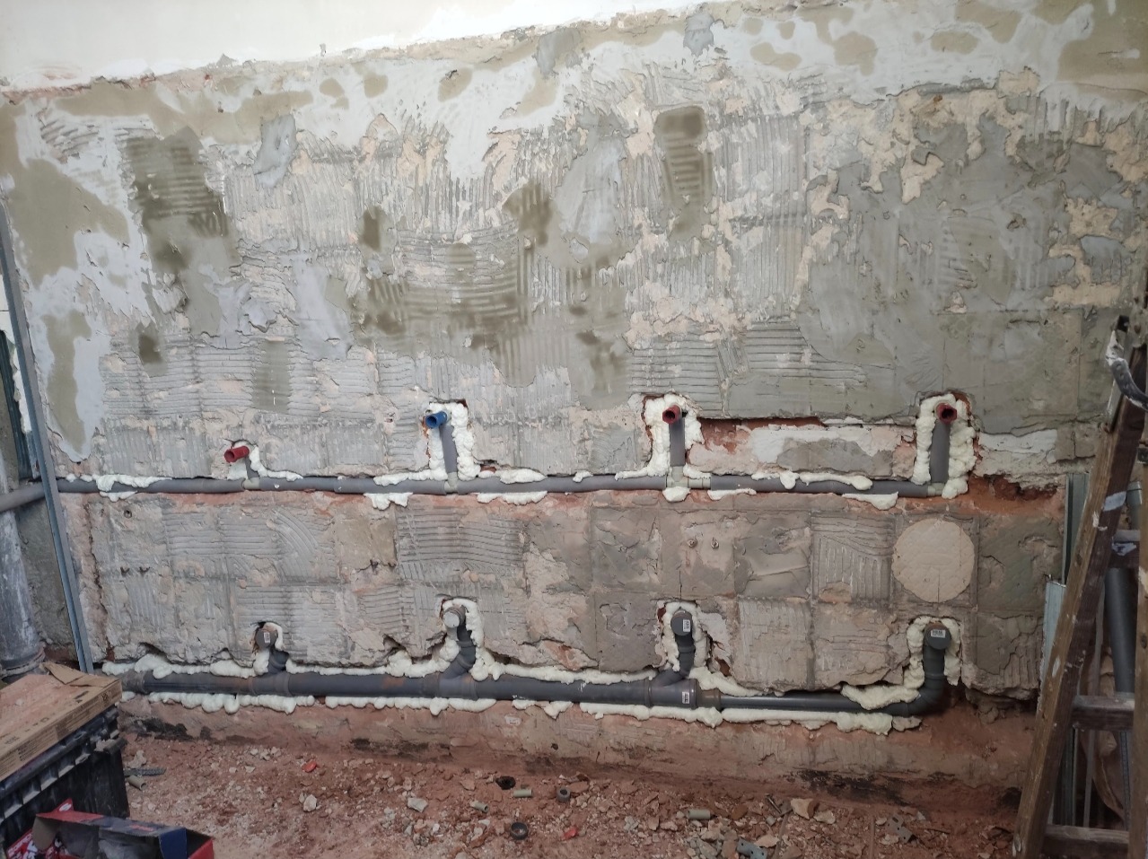 Szare rury instalacji hydraulicznej zamontowane w ścianie z surowego betonu, zabezpieczone pianką montażową. Widoczne fragmenty instalacji z czerwonymi i niebieskimi końcówkami.