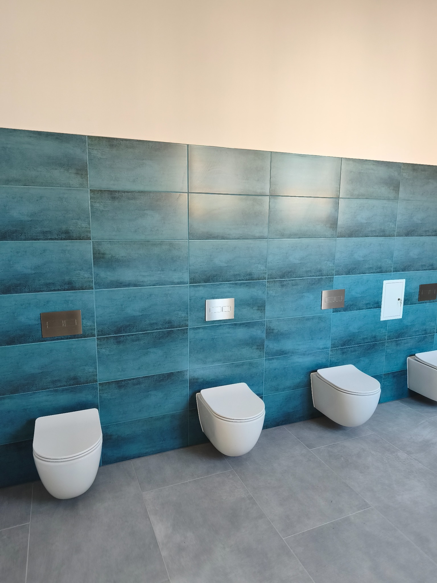 Rząd podwieszanych toalet w nowoczesnej łazience z turkusowymi płytkami i szarą podłogą. Widoczne przyciski spłukujące i rewizja.