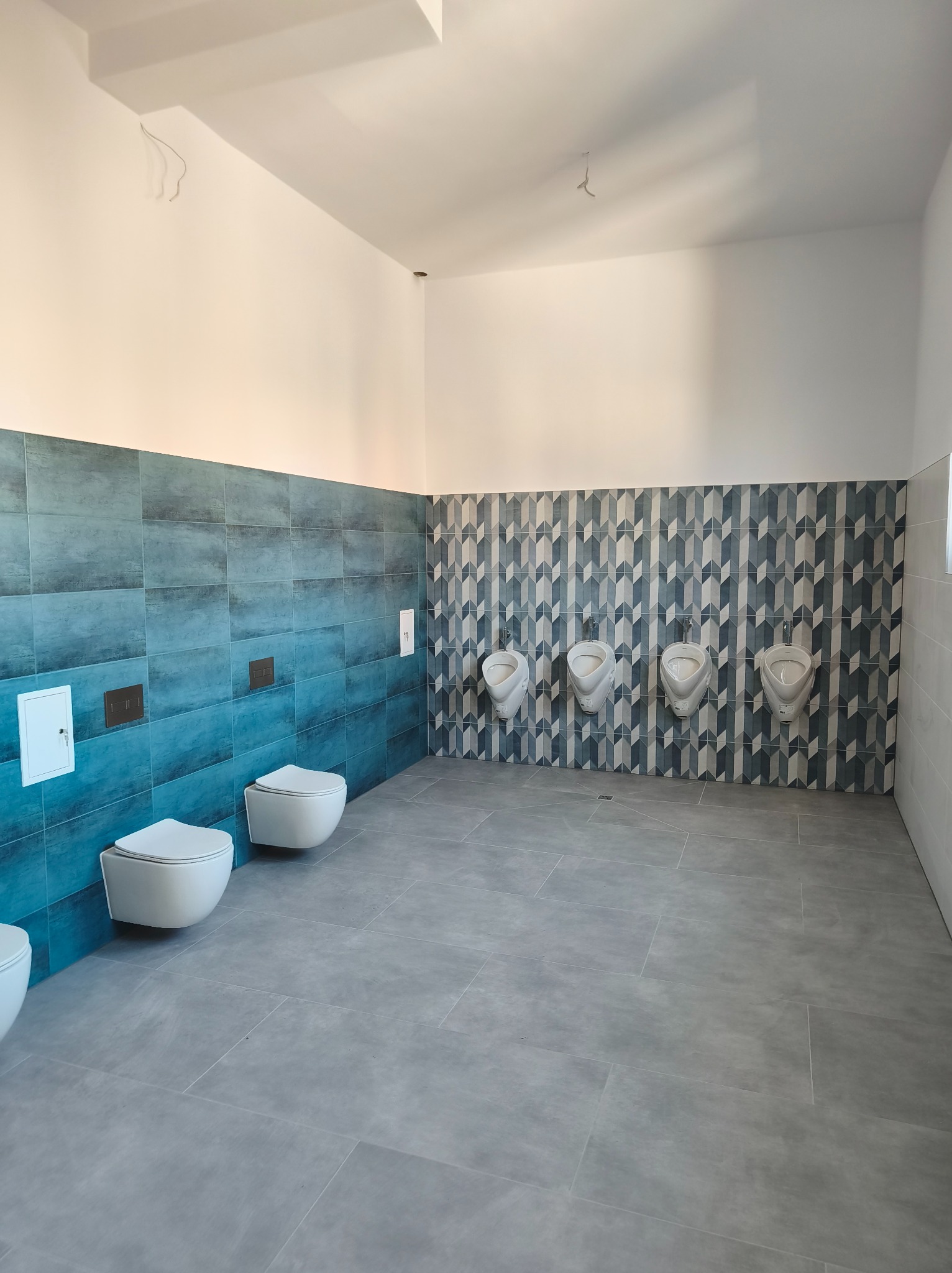 Wykończona łazienka publiczna z czterema pisuarami na ścianie z geometrycznym wzorem i dwoma podwieszanymi toaletami na ścianie z niebieską glazurą.