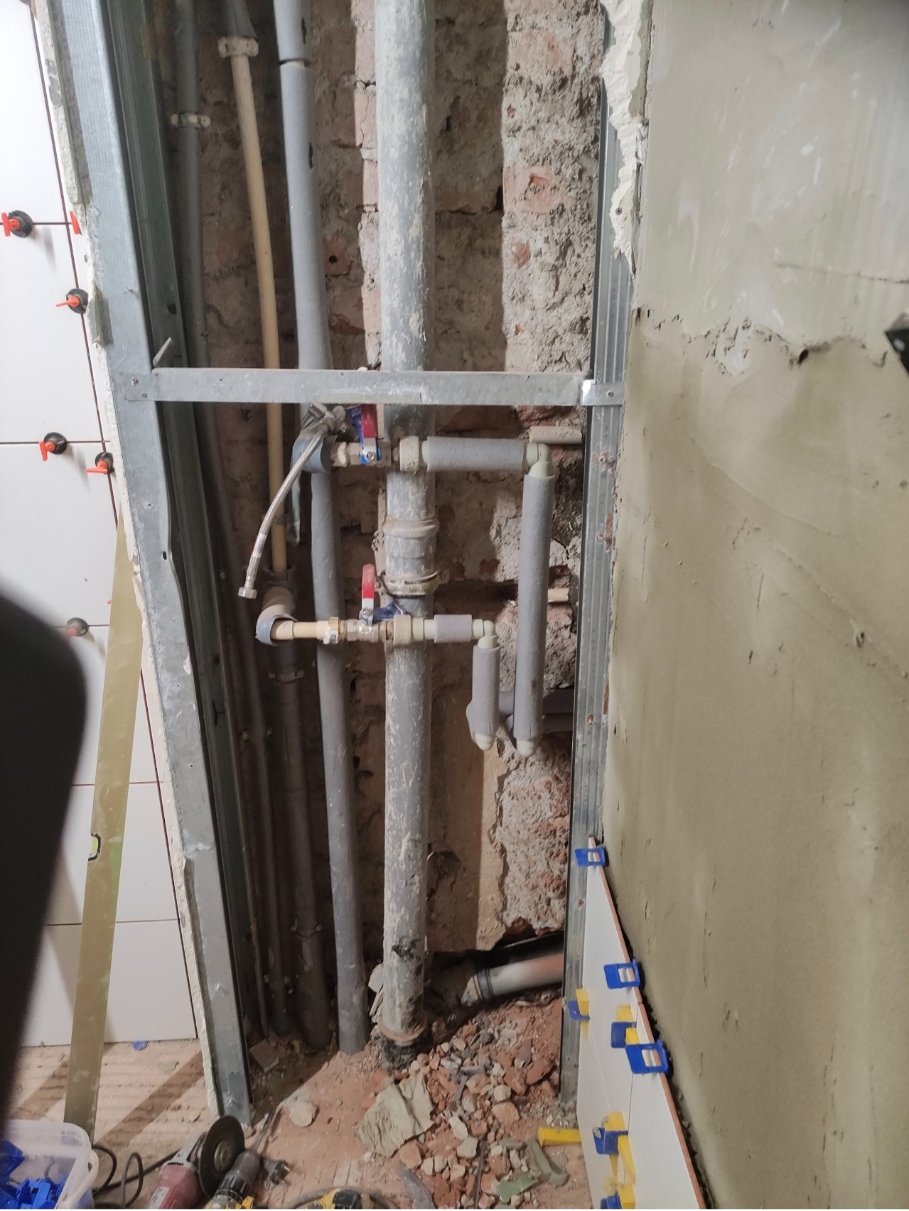 Montaż instalacji wodnej z rurami w szarej izolacji, widoczne zawory i fragment ściany z płytkami. Prace hydrauliczne w trakcie remontu łazienki.