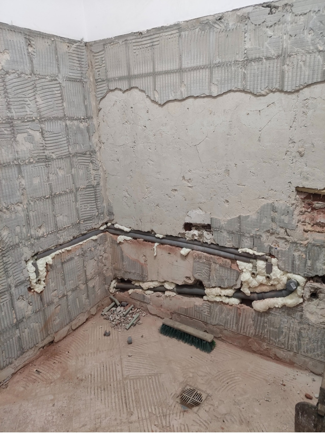 Szare rury hydrauliczne, zabezpieczone pianką montażową, biegną wzdłuż ściany z resztkami płytek w trakcie remontu łazienki. Widoczny fragment podłogi i ściany.