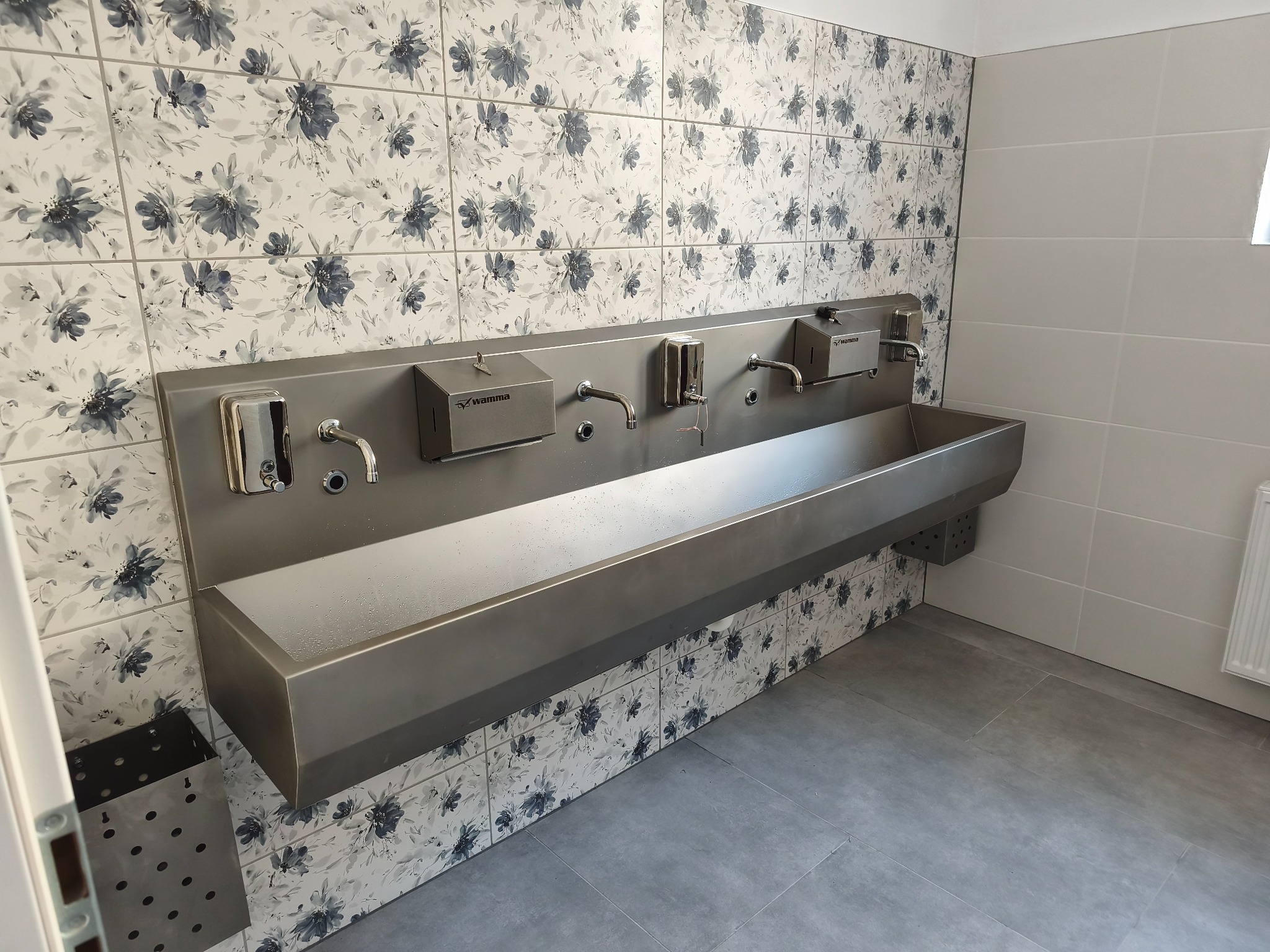 Długa, metalowa umywalka z trzema kranami i dozownikami mydła, zamontowana na ścianie z płytek w kwiatowy wzór. Widoczny odpływ i fragment grzejnika.