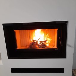 Nowoczesny kominek z czarną obudową i płomieniami. Widoczne drewno w palenisku. Minimalistyczny design, białe ściany. Detal wykończenia wnętrza.