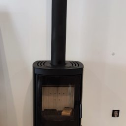 Czarny, wolnostojący kominek z rurą odprowadzającą dym, ustawiony na drewnianej podłodze przy białej ścianie. Wnętrze kominka z widocznym drewnem.