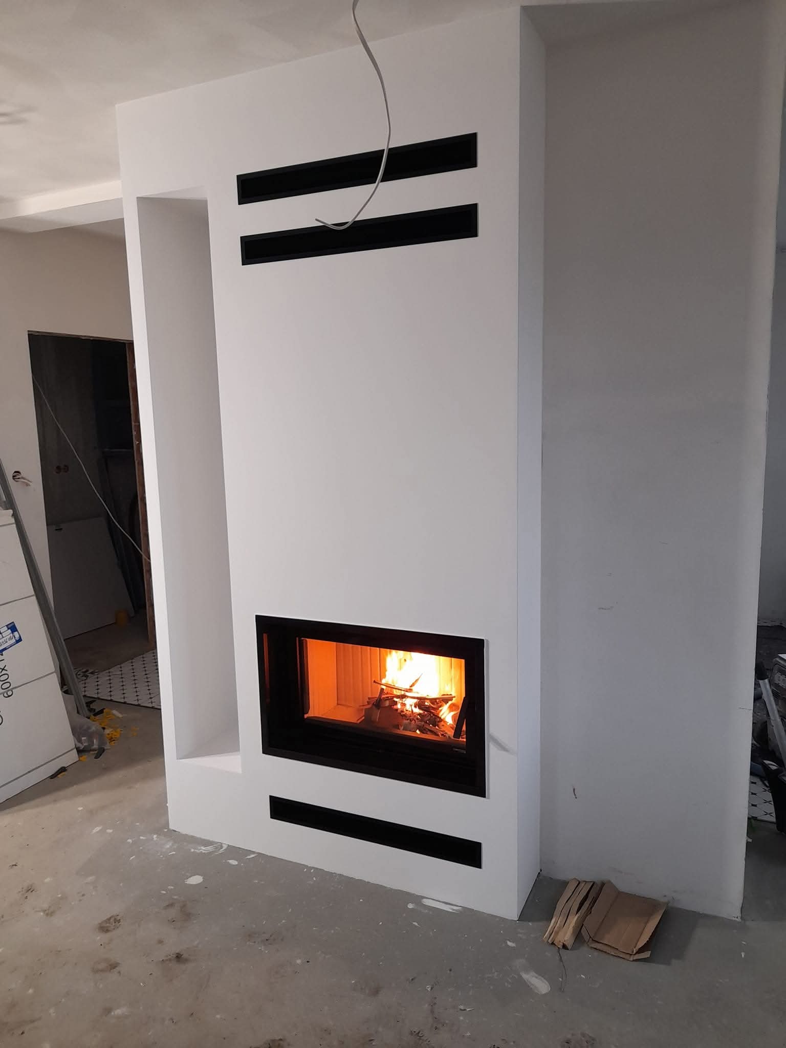 Biały kominek z czarnymi akcentami w nowoczesnym wnętrzu. Widoczne płomienie w palenisku. Surowy stan wykończenia podłogi i ścian. Minimalistyczny design.