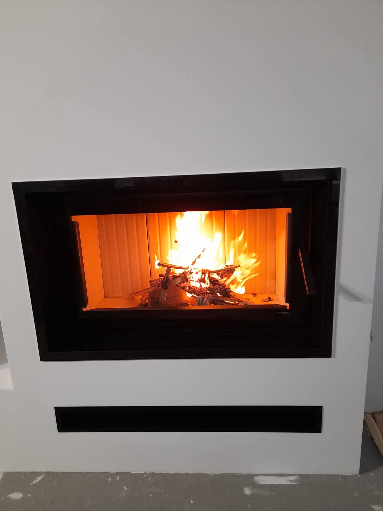 Nowoczesny kominek z czarną obudową i płomieniami. Widoczne drewno w palenisku. Minimalistyczny design, białe ściany. Detal wykończenia wnętrza.