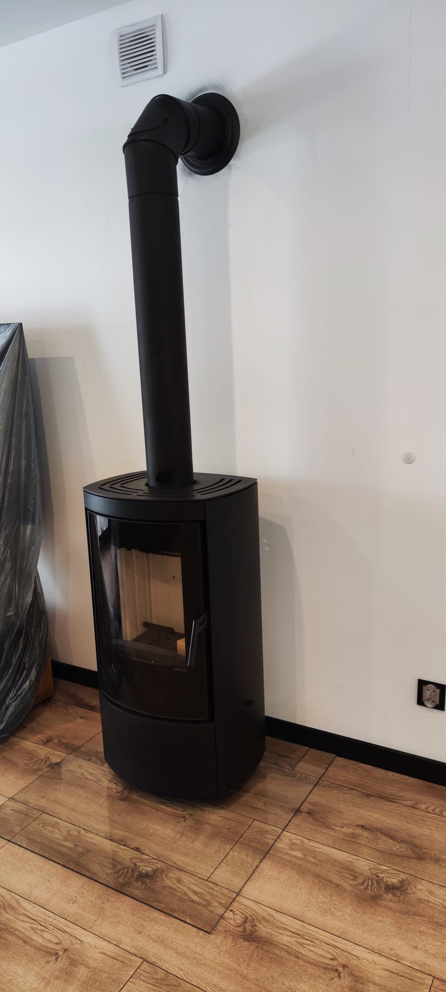 Czarny, wolnostojący kominek z rurą odprowadzającą spaliny, ustawiony na panelach podłogowych. Widoczny fragment ściany z kratką wentylacyjną i gniazdkiem.