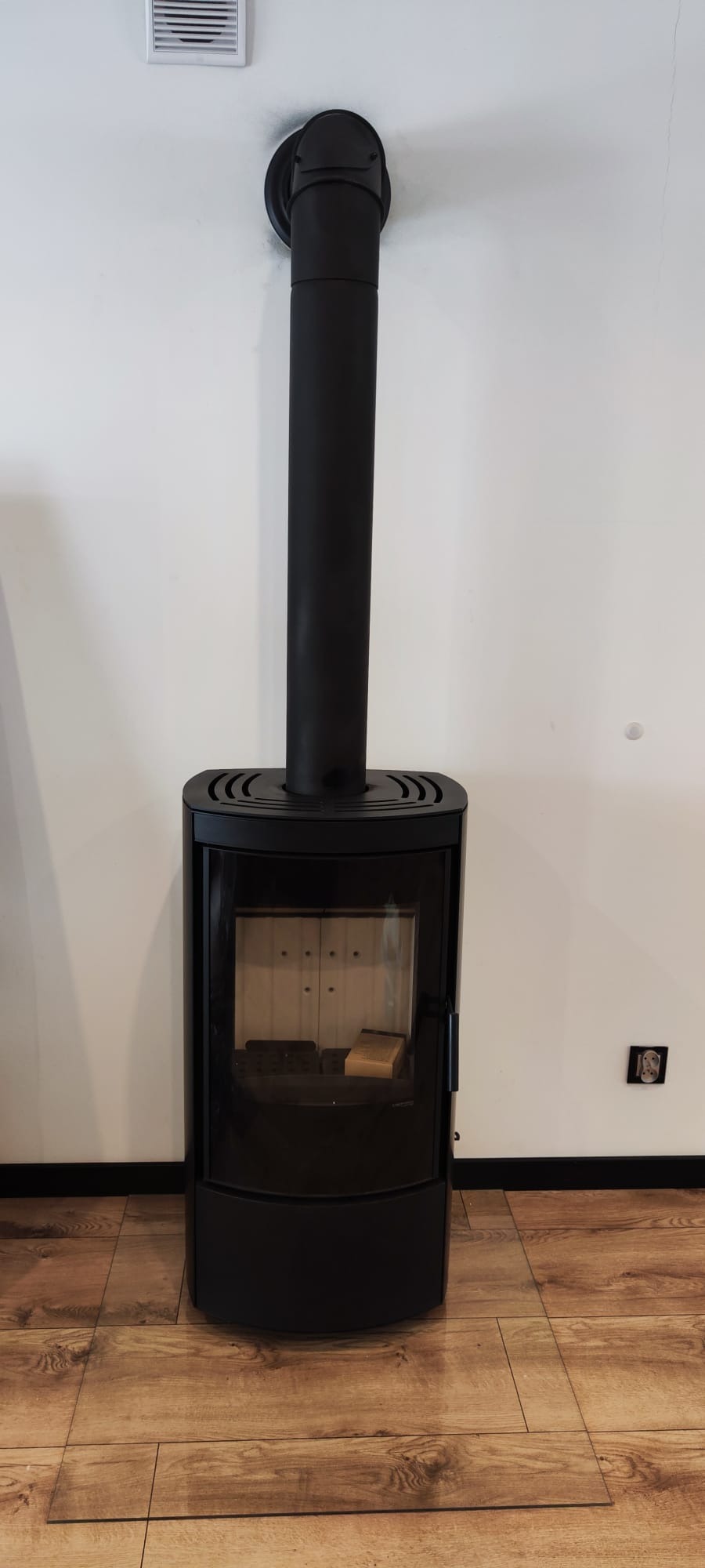 Czarny, wolnostojący kominek z rurą odprowadzającą dym, ustawiony na drewnianej podłodze przy białej ścianie. Wnętrze kominka z widocznym drewnem.