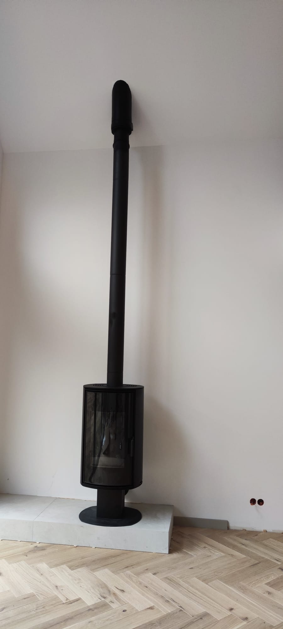 Czarny, nowoczesny kominek wolnostojący z długą rurą odprowadzającą dym, ustawiony na jasnym podeście. Parkiet w jodełkę na podłodze, białe ściany.