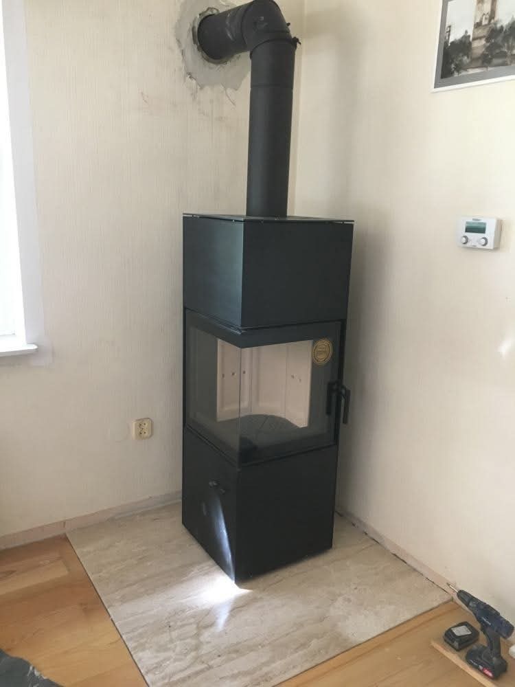 Nowoczesny, czarny kominek wolnostojący z przeszklonym paleniskiem, zainstalowany w rogu pokoju na marmurowej płycie. Widoczny przewód kominowy i narzędzia.