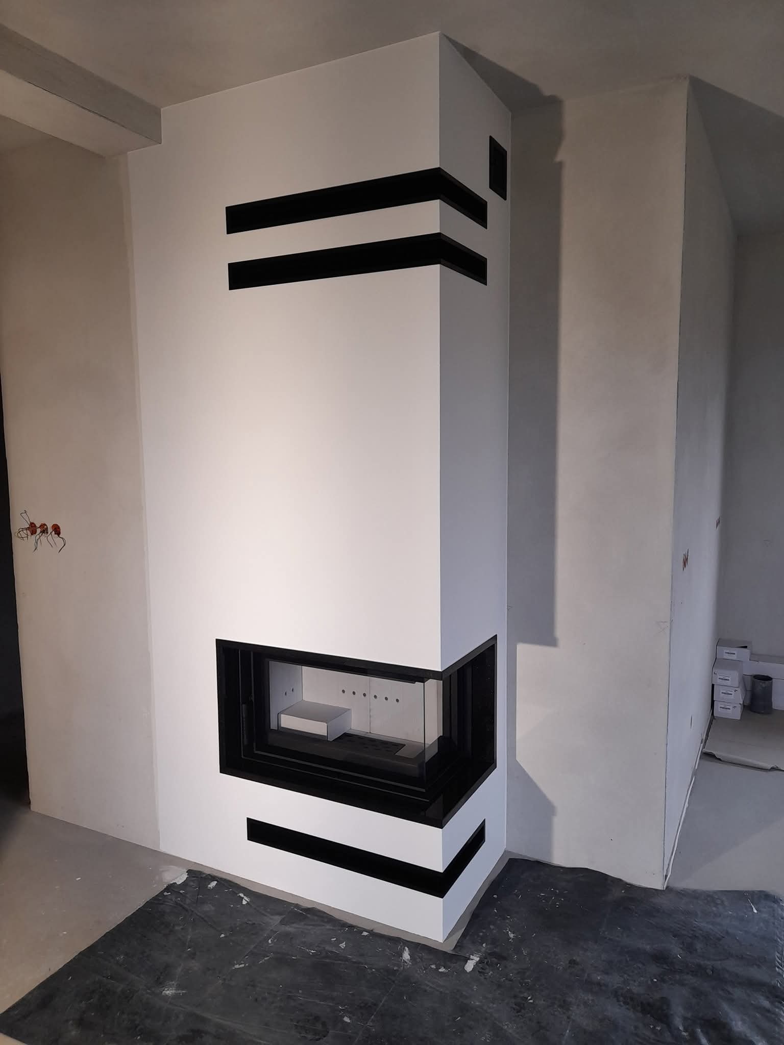 Biały kominek narożny z czarnymi, poziomymi akcentami w surowym wnętrzu. Widoczne niedoskonałości wykończenia i zabezpieczenie podłogi folią malarską.