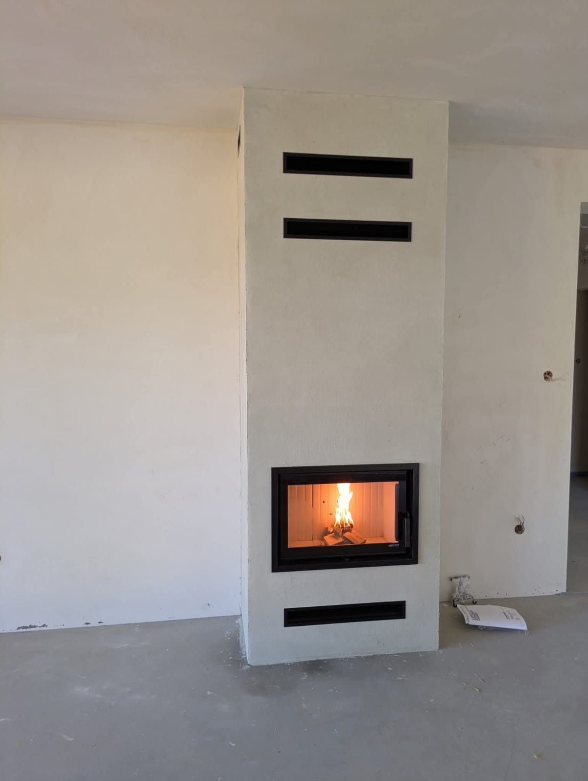 Nowoczesny kominek z czarną obudową i płomieniem, w surowym wnętrzu z jasnymi ścianami i betonową podłogą. Widoczne czarne poziome wloty powietrza.