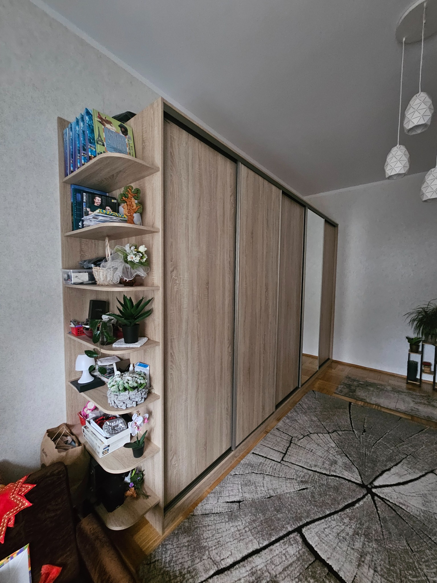 Jasna szafa przesuwna z półkami narożnymi, wypełniona dekoracjami i książkami. Na podłodze dywan imitujący przekrój pnia drzewa. Widoczne lampy wiszące.