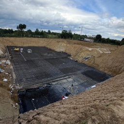 BRAVEX TARAS PAVLIUK - Szeroki wykop pod fundamenty z ułożoną siatką zbrojeniową na czarnej folii. W tle widoczne drzewa i niebo z chmurami. Na górze wykopu stoi mała koparka.