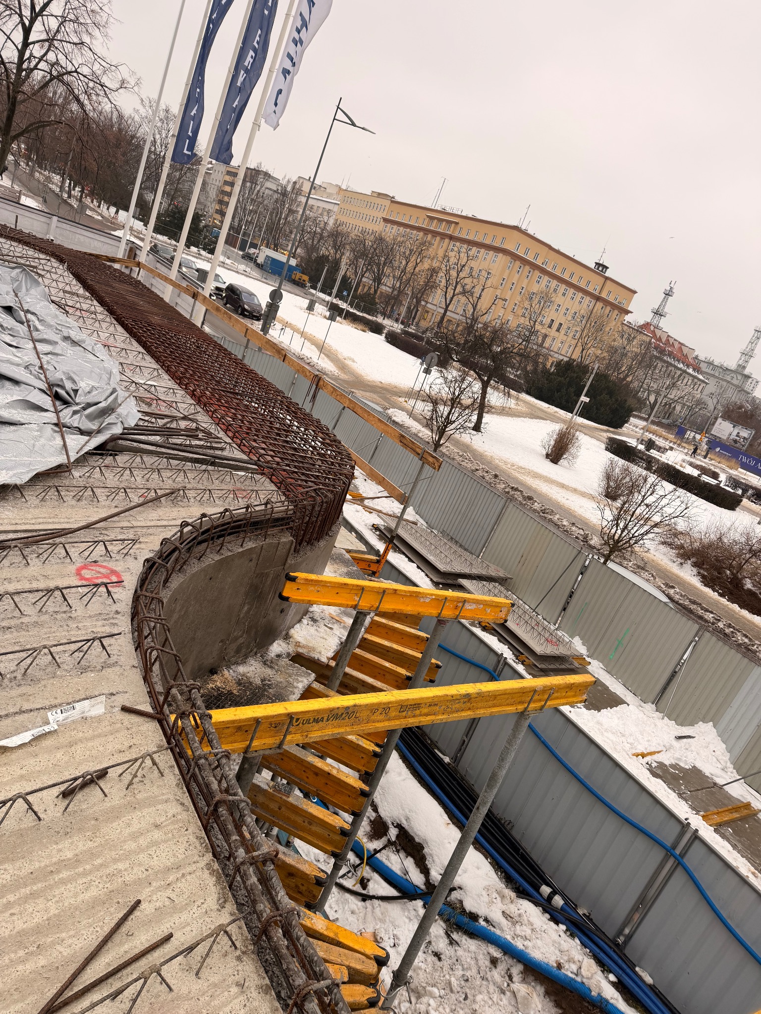 Zbrojenie fundamentów na budowie w zimowej scenerii. Widoczne stalowe pręty, szalunki i elementy konstrukcyjne na tle zaśnieżonego terenu miejskiego.