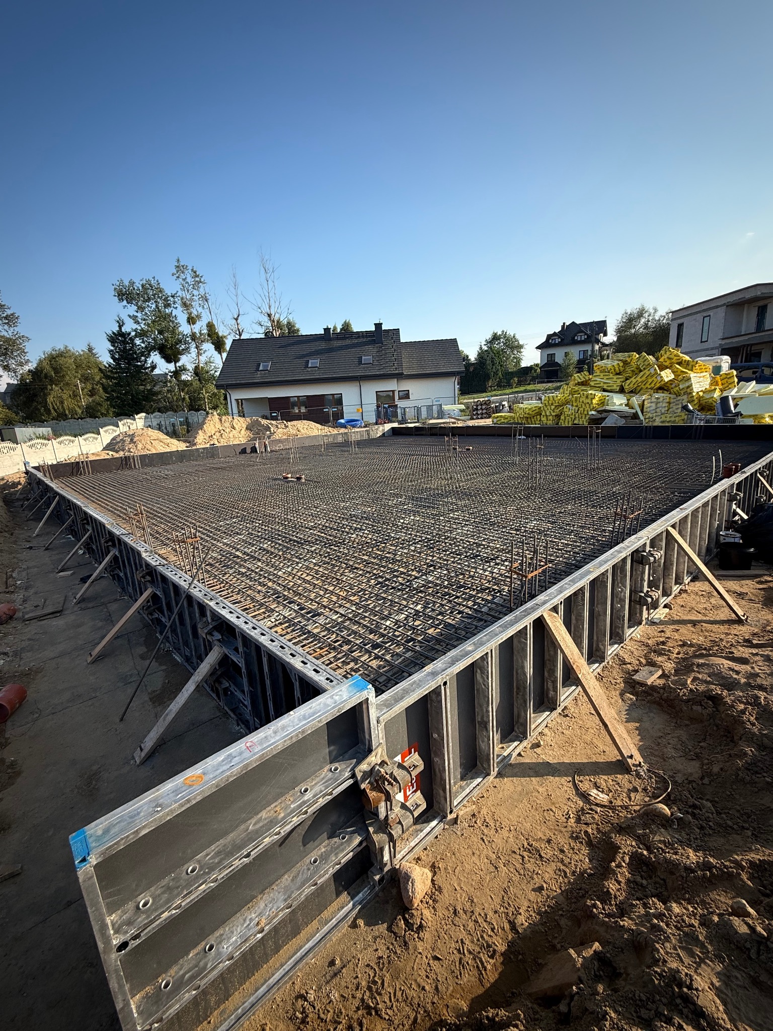 Zbrojenie fundamentu domu jednorodzinnego, widok z góry na siatkę ze stali, szalunki z metalu, w tle budynki i niebo. Prace budowlane w toku.
