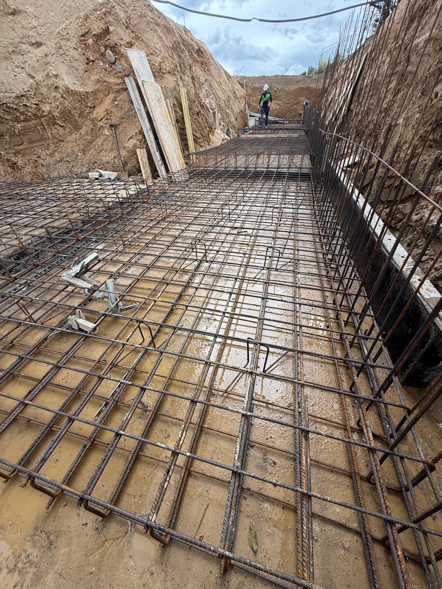 Zbrojenie fundamentów w wykopie, widoczne stalowe pręty tworzące siatkę, częściowo zalane wodą. W tle pracownik w kasku i kamizelce odblaskowej.