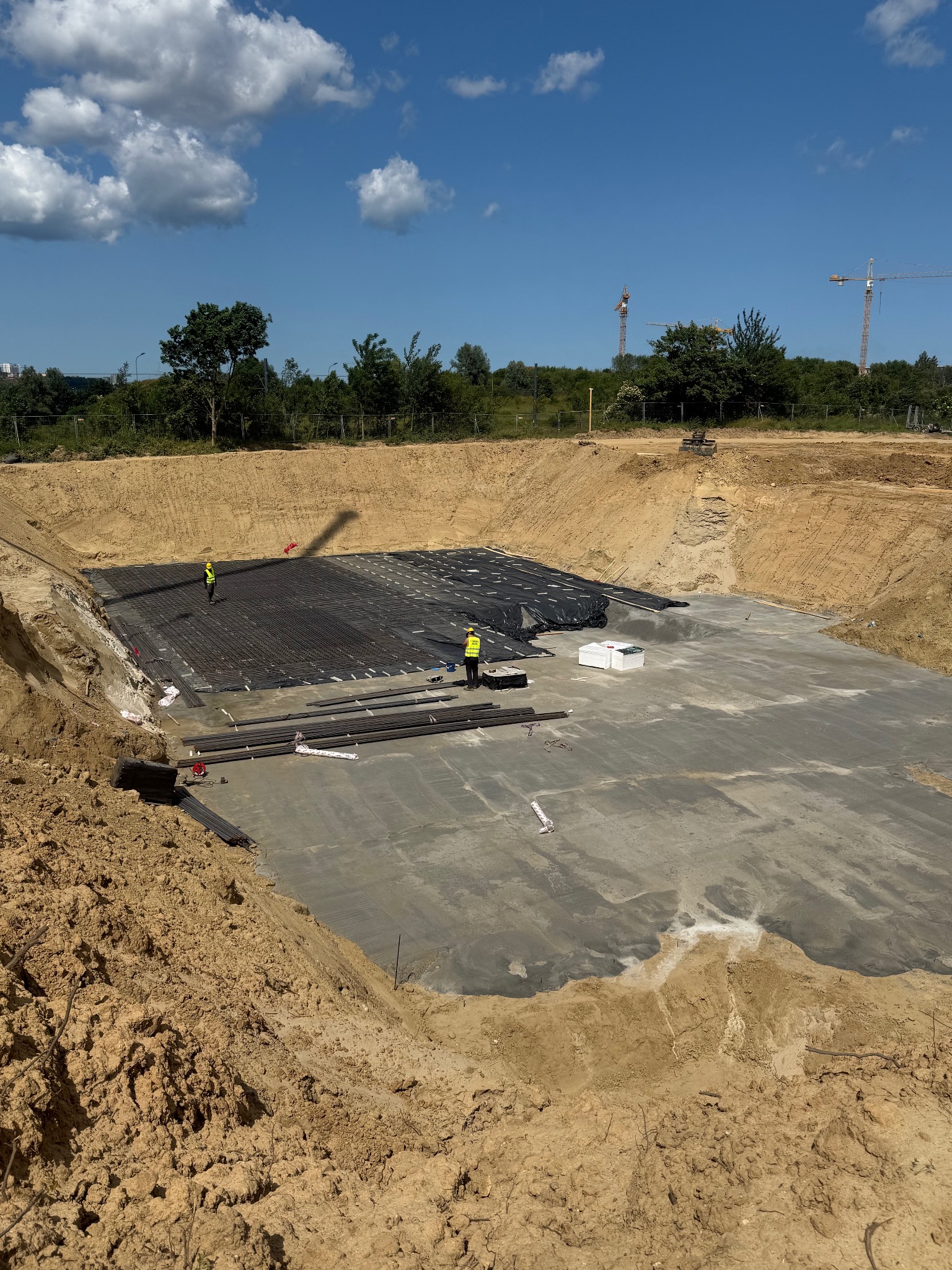 Przygotowanie fundamentów pod budynek w Gdyni. Widoczne zbrojenie i pracownicy w żółtych kamizelkach na placu budowy. Ujęcie z góry, słoneczny dzień.
