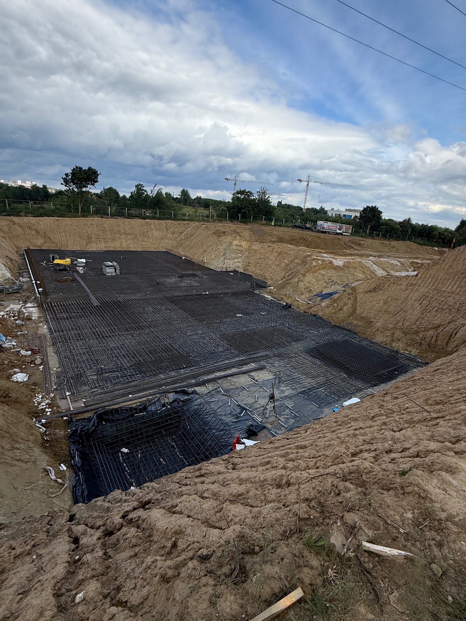 Szeroki wykop pod fundamenty z ułożoną siatką zbrojeniową na czarnej folii. W tle widoczne drzewa i niebo z chmurami. Na górze wykopu stoi mała koparka.