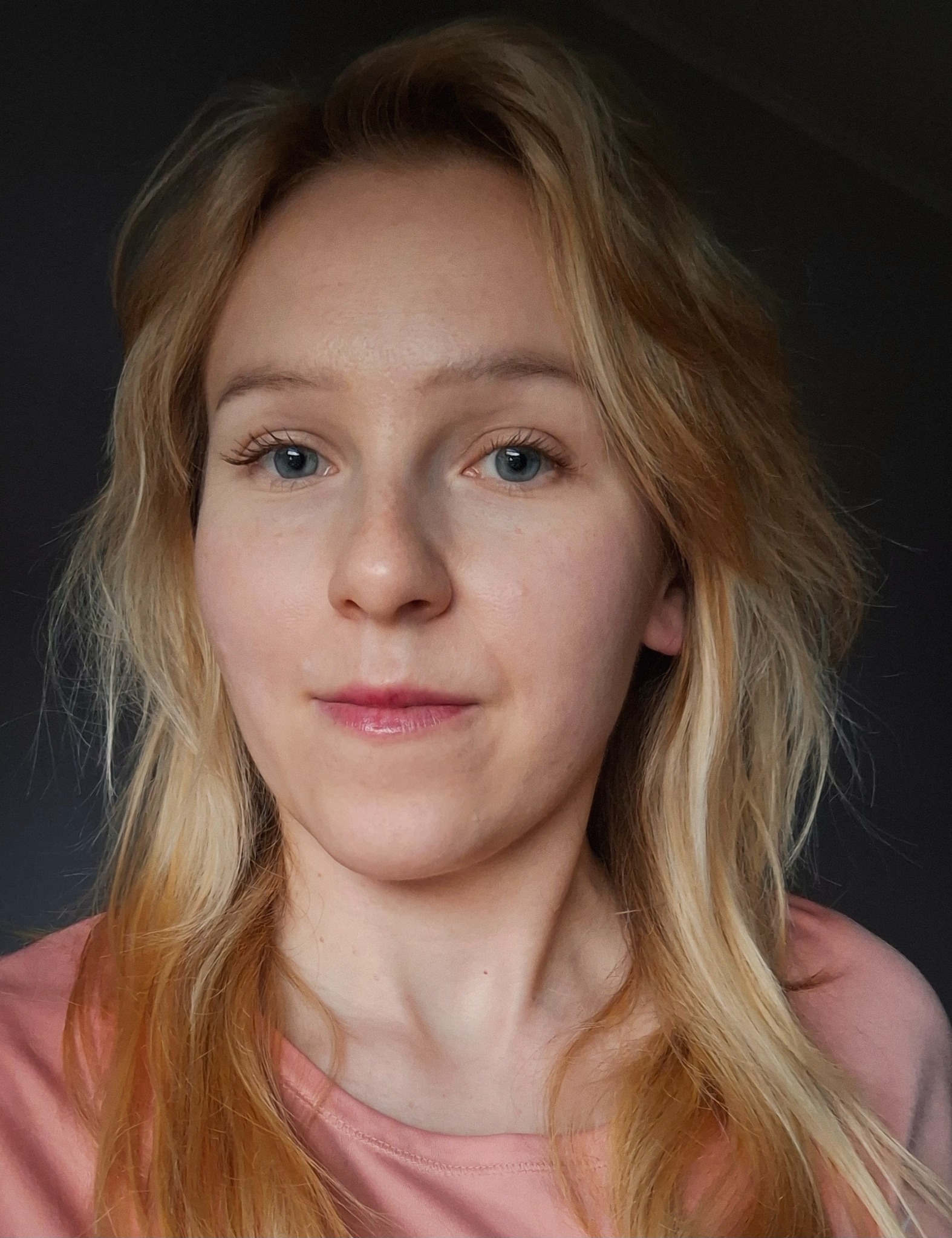 Portret kobiety o jasnych włosach i niebieskich oczach, ubrana w różową koszulkę, na ciemnym tle. Naturalny makijaż, delikatny uśmiech.