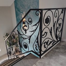 DAM-DAR METAL DAMIAN KRZEMIEŃ - Kuta, czarna balustrada schodowa z dekoracyjnym motywem roślinnym, widziana z góry. W tle ściana z efektem ombre i okno z rośliną. Szare płytki podłogowe.