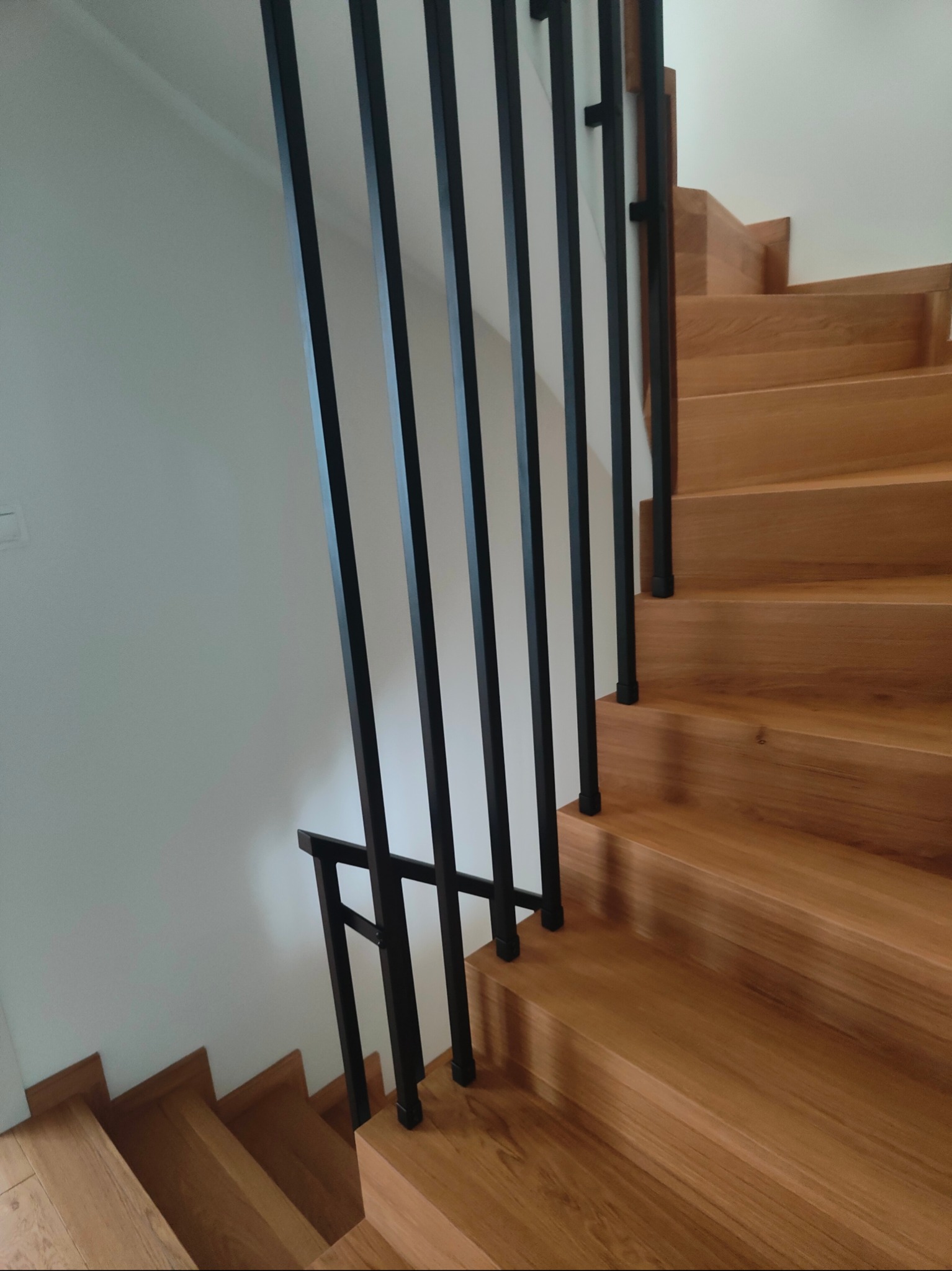 Nowoczesne schody drewniane z czarną, minimalistyczną balustradą metalową. Jasne drewno stopni kontrastuje z ciemnym metalem. Ujęcie z dołu, podkreślające geometrię.