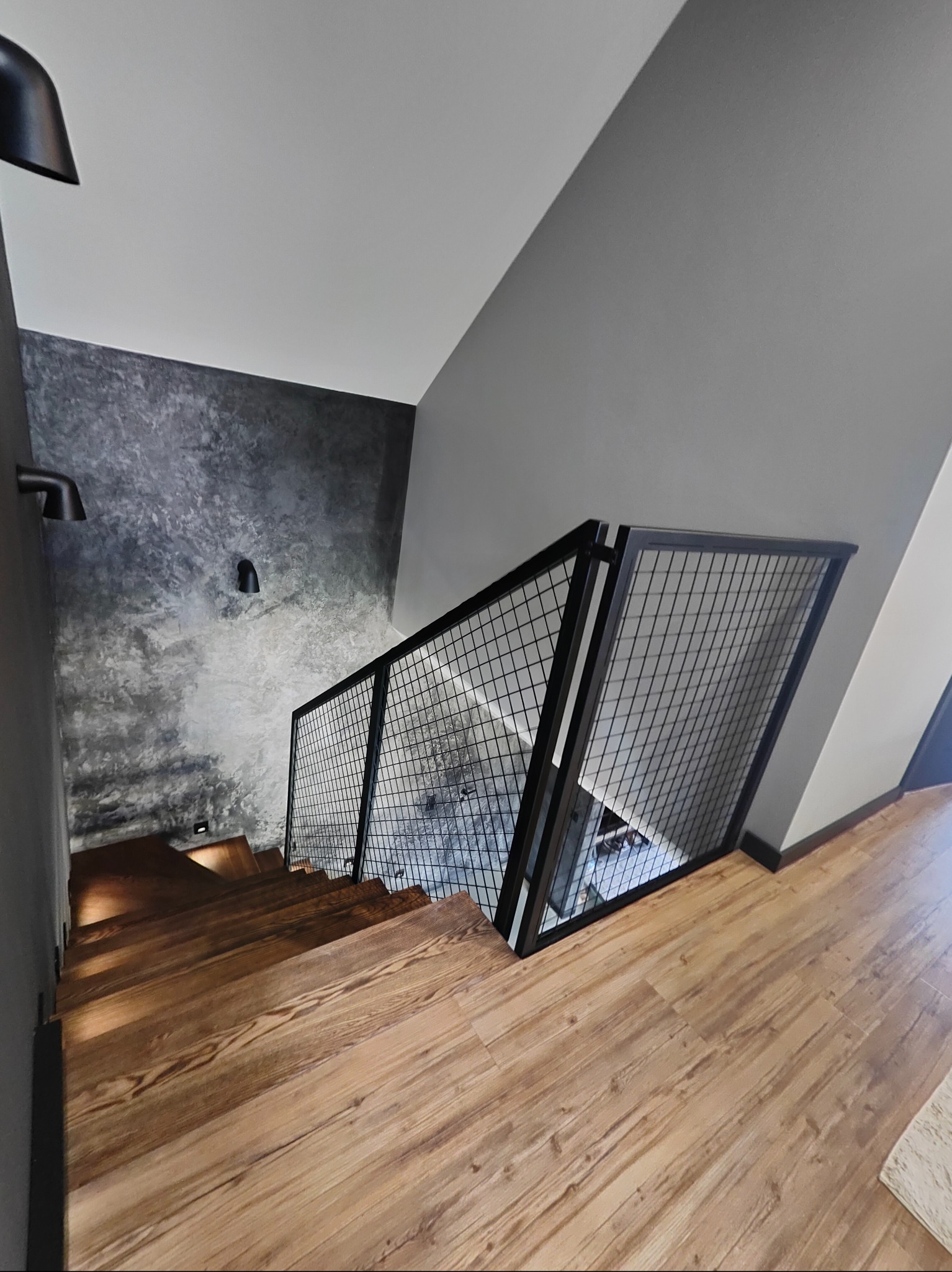 Nowoczesne schody drewniane z czarną, metalową balustradą z siatki, oświetlone punktowo, na tle szarej ściany o fakturze betonu. Widok z góry.