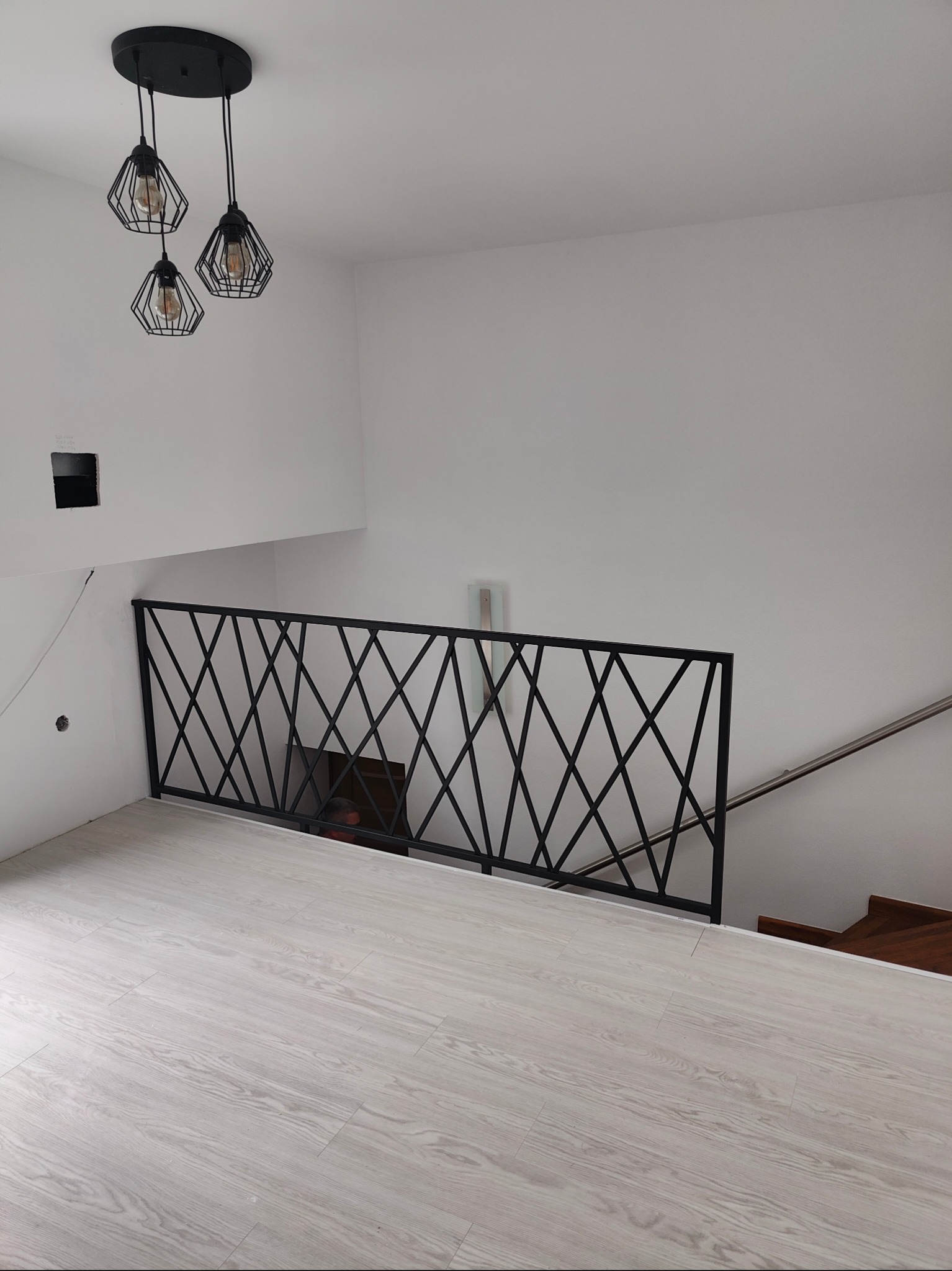 Nowoczesna, czarna balustrada metalowa z geometrycznym wzorem krzyżowym, zamontowana na antresoli z drewnianymi schodami. Minimalistyczne oświetlenie.