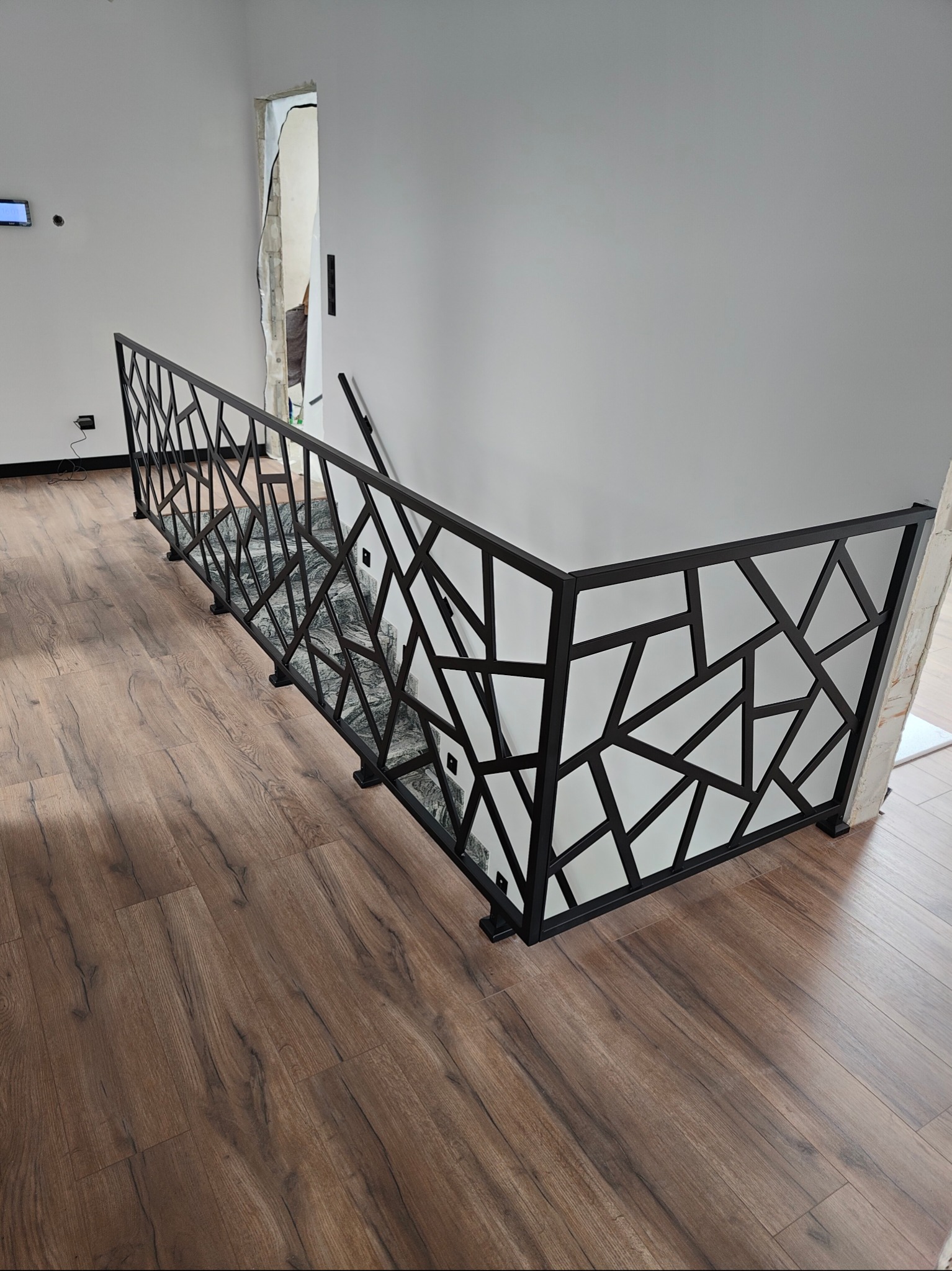 Czarna, geometryczna balustrada schodowa z metalu, zamontowana w nowoczesnym wnętrzu z panelami podłogowymi. Widok z góry na ażurową konstrukcję.