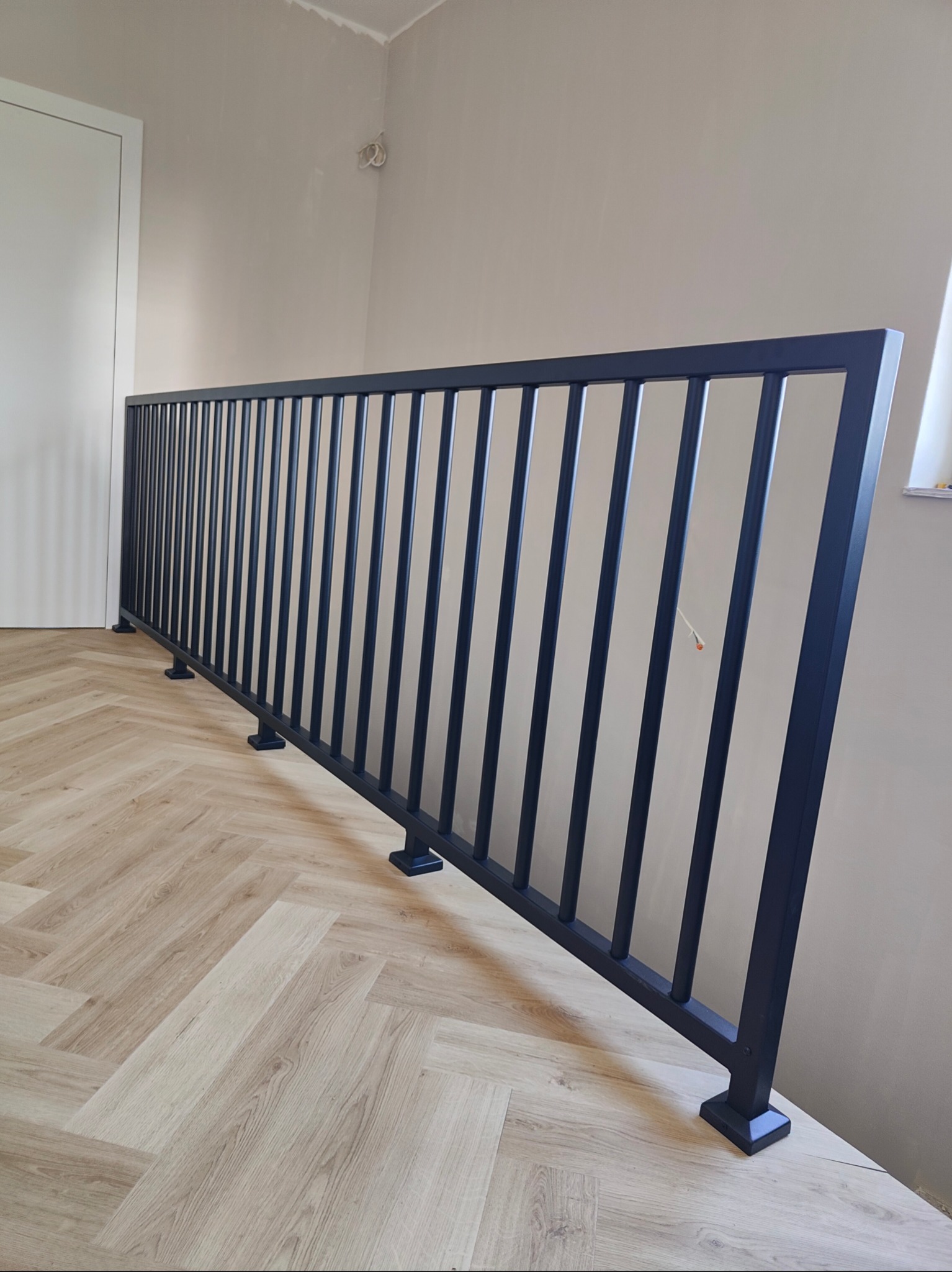 Nowoczesna, czarna balustrada schodowa z pionowymi szczeblami, zamontowana na jasnej podłodze w jodełkę. Minimalistyczny design, widok z boku, neutralne tło.